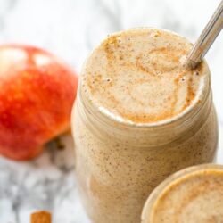 Two apple cinnamon smoothies in mason jars next to a red apple and cinnamon sticks
