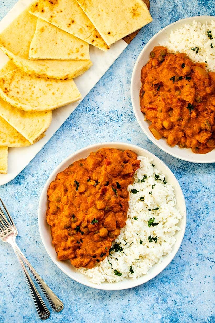 Two servings of potato and chickpea tikka masala with basmati rice on white plates next to sliced naan on a serving platter