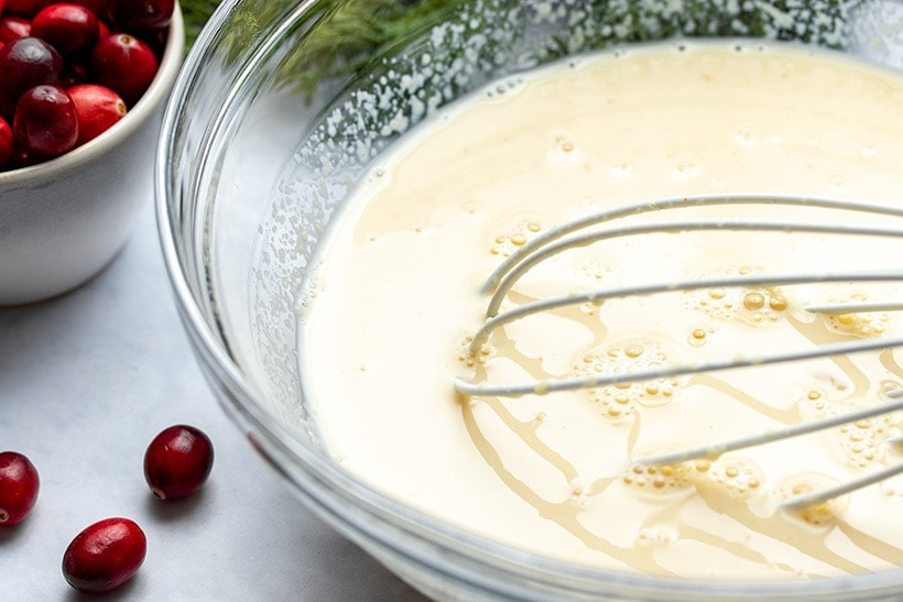 Milk, applesauce, egg and vanilla being whisked in a glass bowl next to a measuring cup filled with cranberries