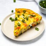 A slice of potato and spinach frittata on a cream coloured plate with a small blue bowl of diced green onion in the background
