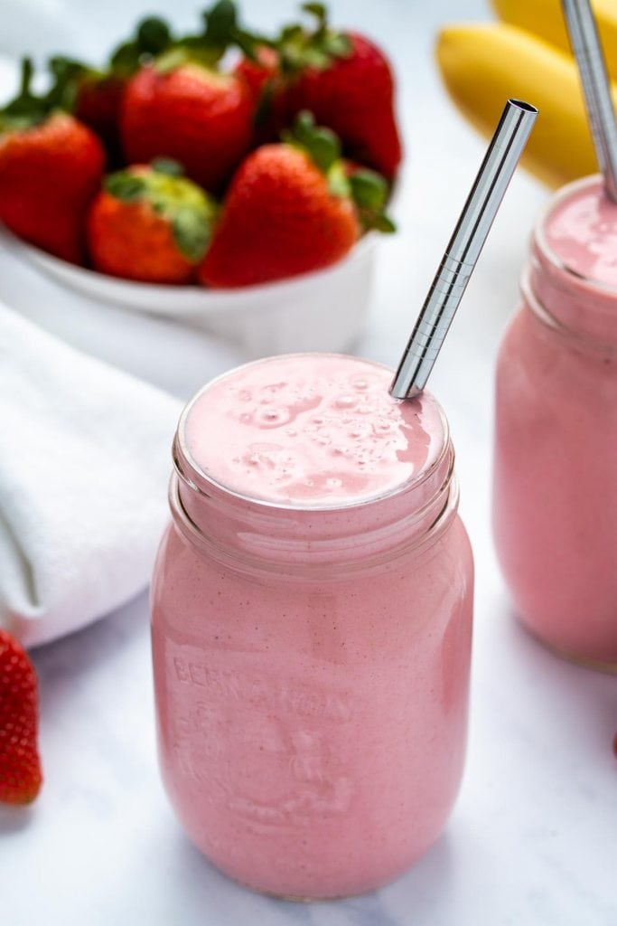 two strawberry banana smoothies in mason jars with stainless steel straws in front of a white bowl of strawberries and a bunch of bananas