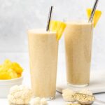 Two glasses of cauliflower smoothie garnished with pineapple and a silver straw surrounded by cauliflower florets, hemp hearts and a white bowl of diced pineapple