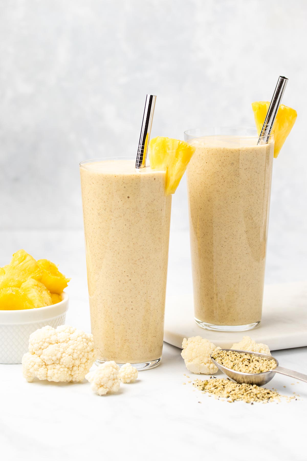 Two glasses of cauliflower smoothie garnished with pineapple and a silver straw surrounded by cauliflower florets, hemp hearts and a white bowl of diced pineapple