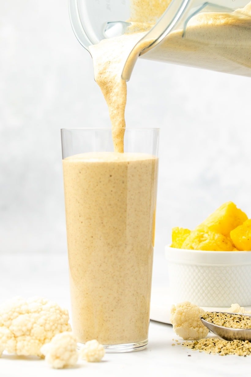 Cauliflower smoothie being poured from a blender into a glass, surrounded by cauliflower florets, hemp hearts and a white bowl of diced pineapple