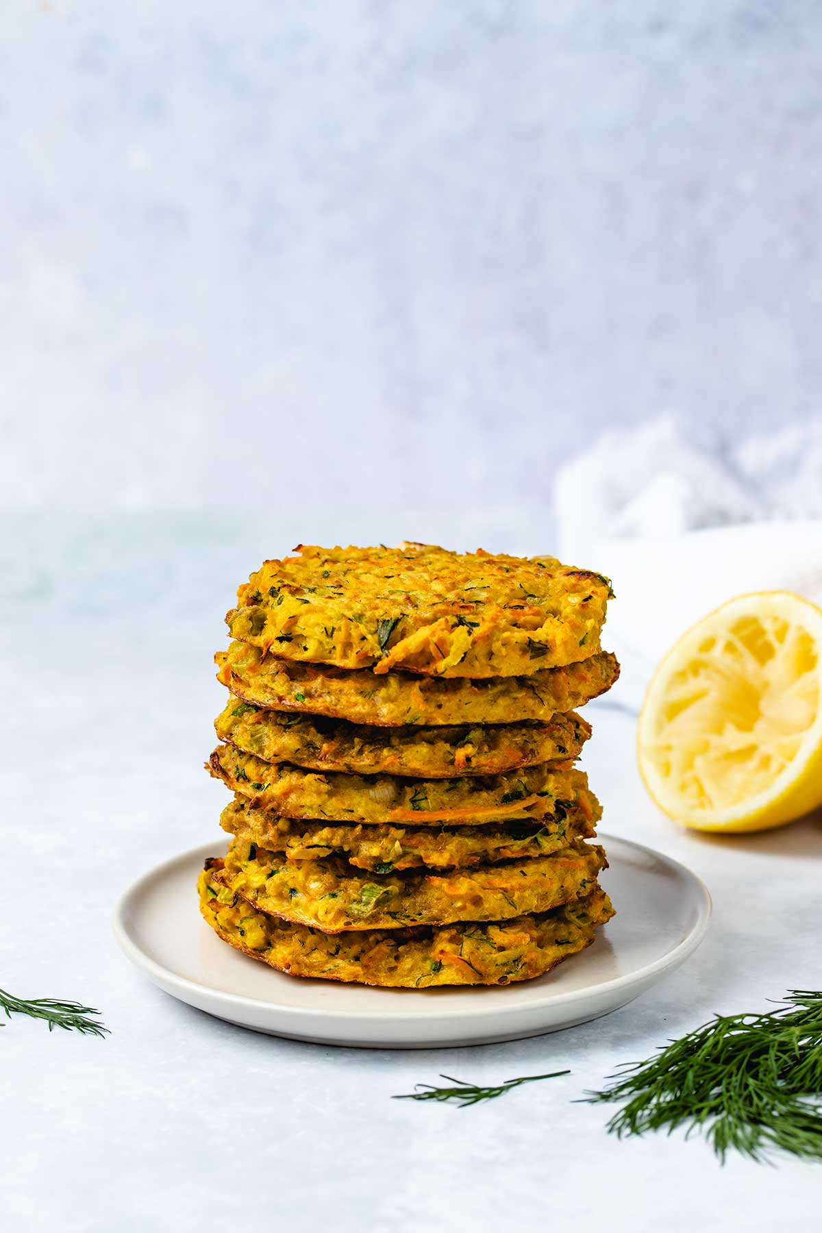 A stack of 7 zucchini carrot fritters on a cream coloured plate next to dill garnish and half a squeezed lemon