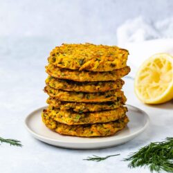 A stack of 7 zucchini carrot fritters on a cream coloured plate next to dill garnish and half a squeezed lemon