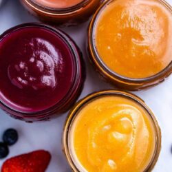 Four flavours of Instant Pot applesauce in decorative mason jars surrounded by strawberries, blueberries and a peach