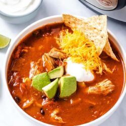 chicken enchilada soup in a white bowl garnished with diced avocado, sour cream, grated cheese and corn chips, sitting in front of an Instant Pot