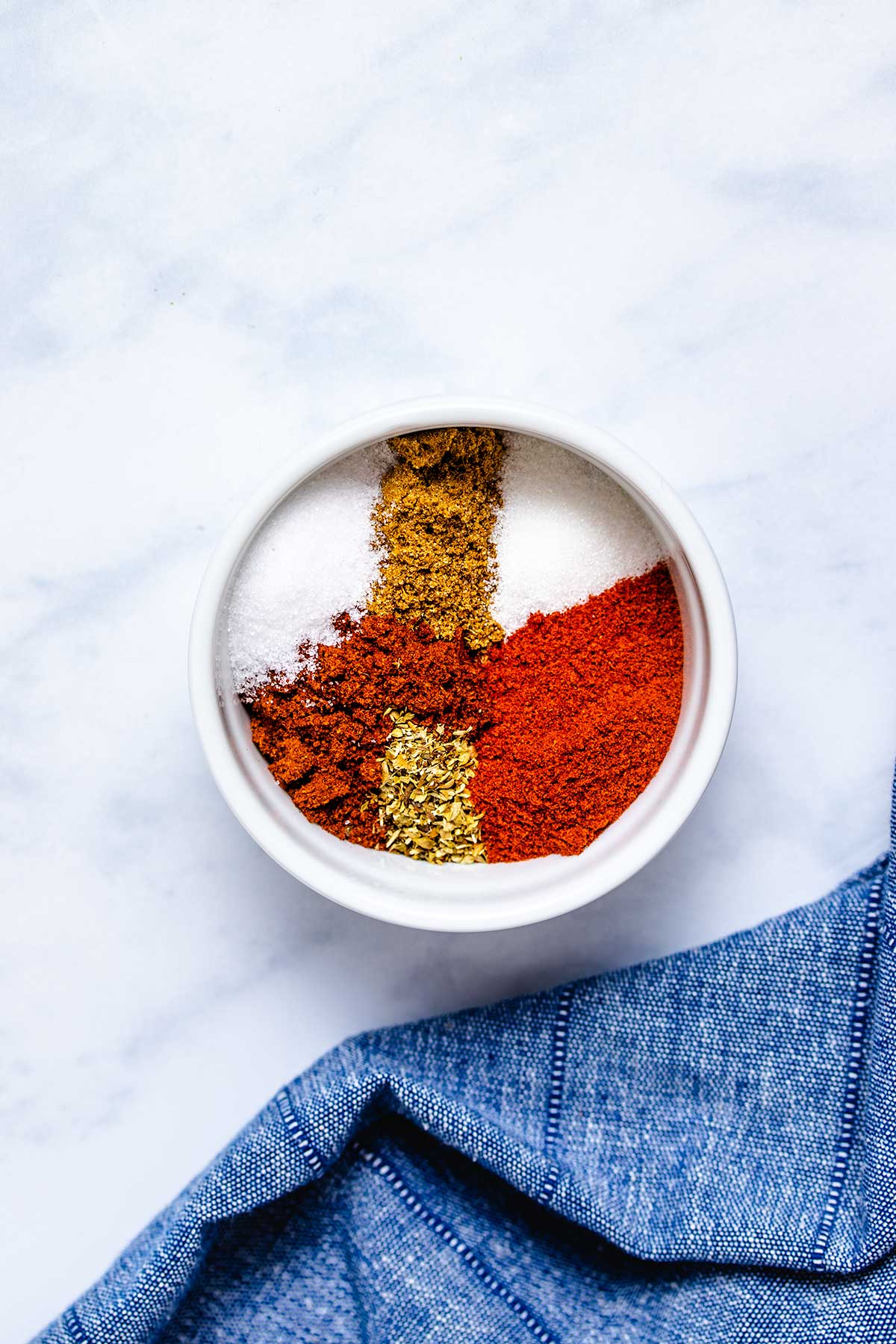 Chili powder, smoked paprika, cumin, oregano, salt and sugar in a small white bowl next to a denim linen