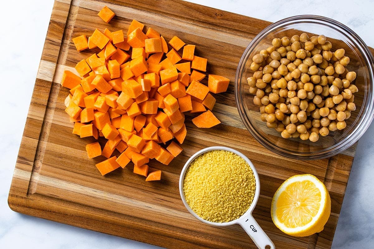 diced sweet potato, a bowl of chickpeas, a measuring cup of uncooked couscous and half a lemon on a wooden cutting board