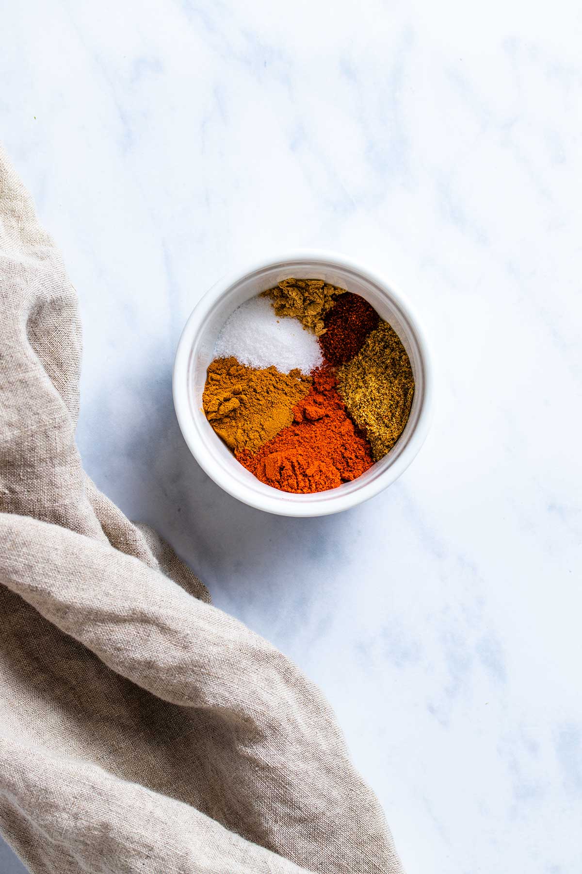 moroccan spices in a small white bowl next to a light brown linen with a white marble background