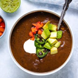 black bean soup in a white bowl garnished with sour cream, sliced jalapeno, diced red pepper and diced avocado, with a spoon inserted