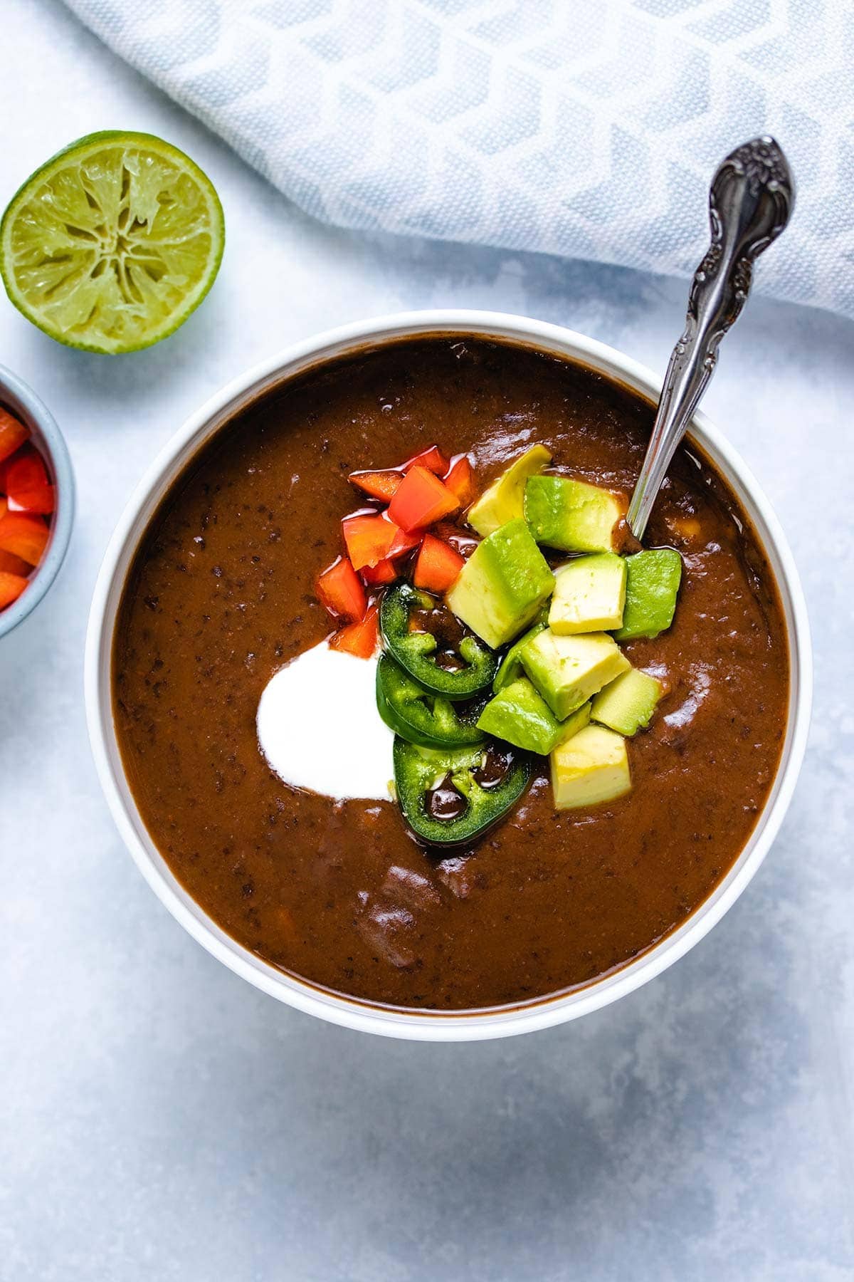 black bean soup in a white bowl garnished with sour cream, sliced jalapeno, diced red pepper and diced avocado, with a spoon inserted