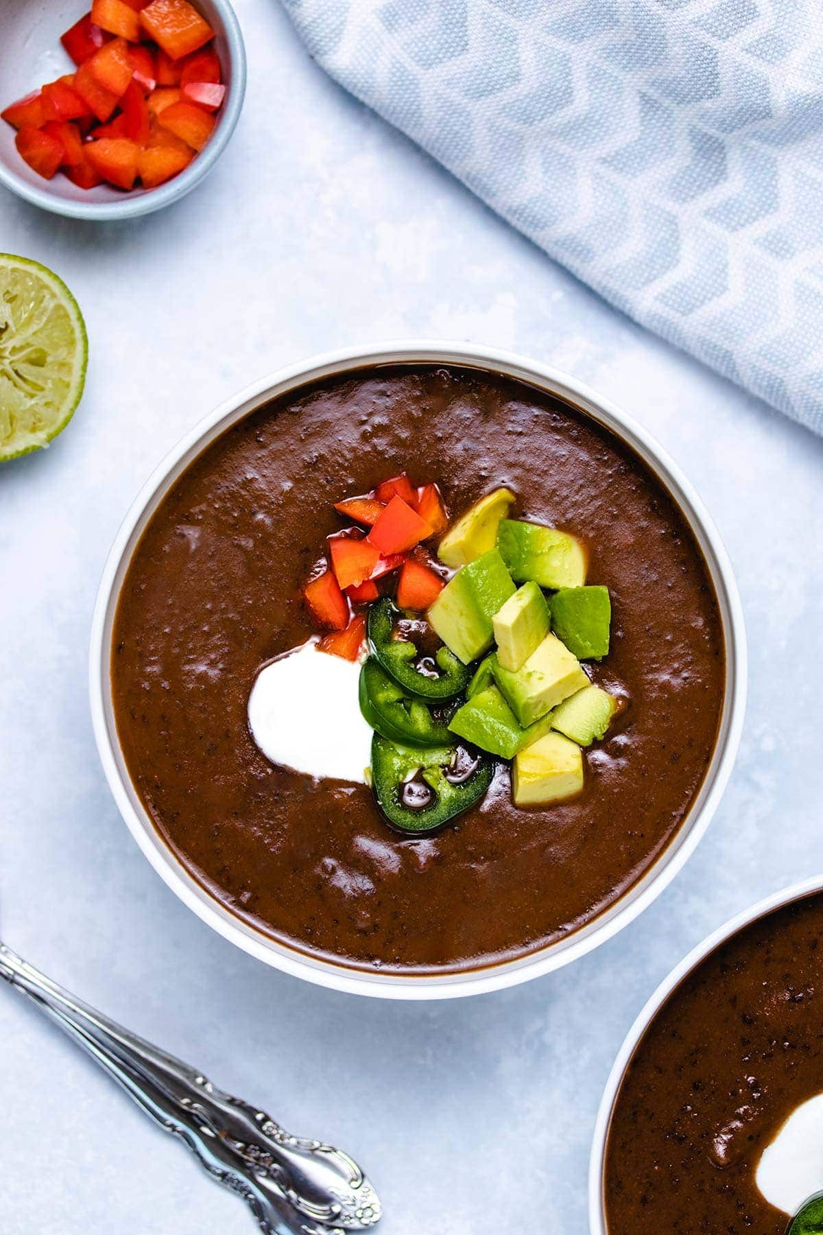 black bean soup in a white bowl garnished with sour cream, sliced jalapeno, diced red pepper and diced avocado