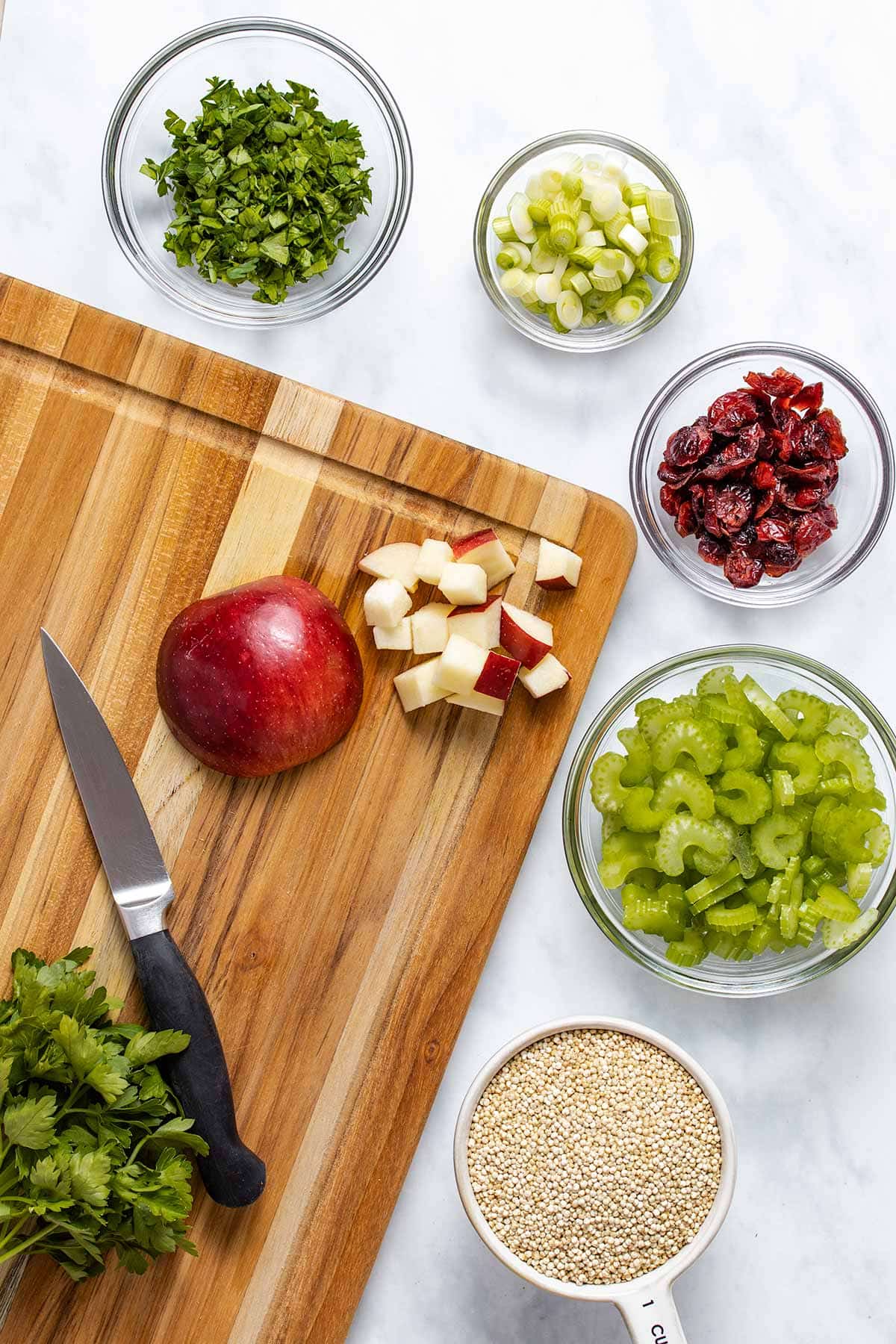 diced apple on a wooden cutting board with a knife, next to small bowls of uncooked quinoa, diced celery, dried cranberries, sliced green onion and chopped parsley