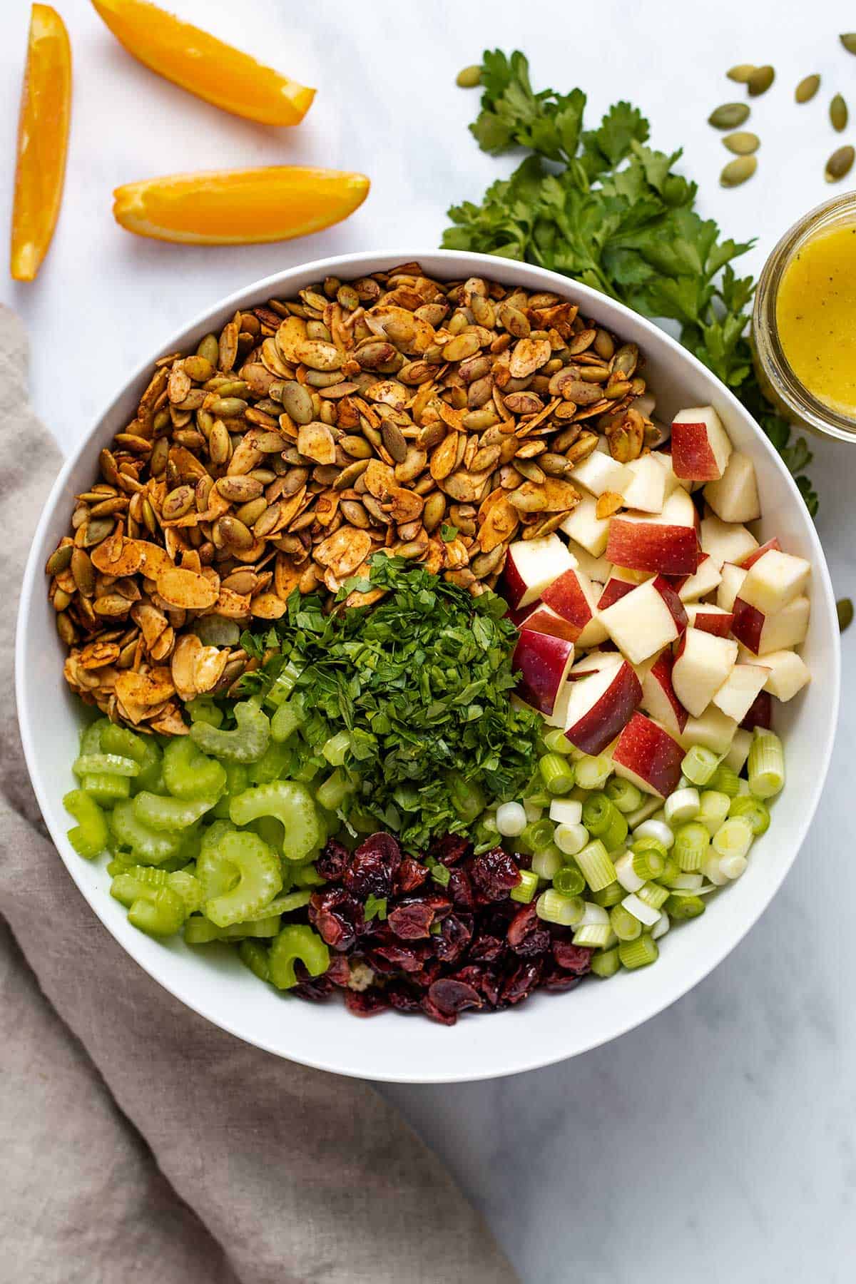 diced apple, sliced green onion, dried cranberries, chopped parsley, diced celery, roasted almond and pumpkin seeds in a white bowl viewed from overhead, next to a mason jar of dressing, a bunch of parsley and 3 orange slices