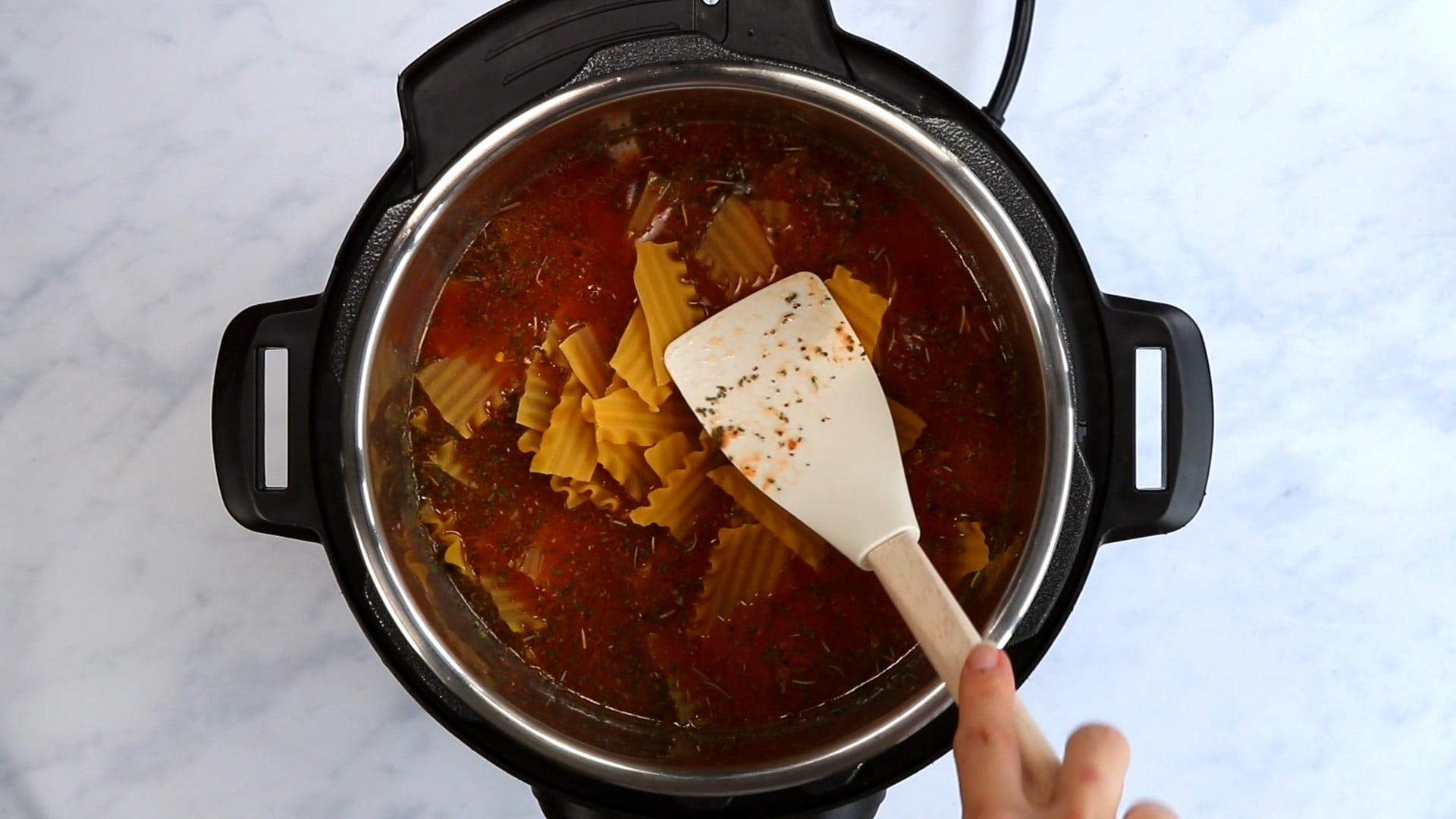 A white spatula pushing broken lasagna noodles into broth in an Instant Pot