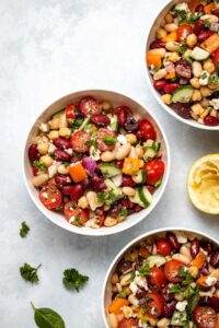Mediterranean Bean Salad in 3 white bowls next to a squeezed lemon half and fresh herbs