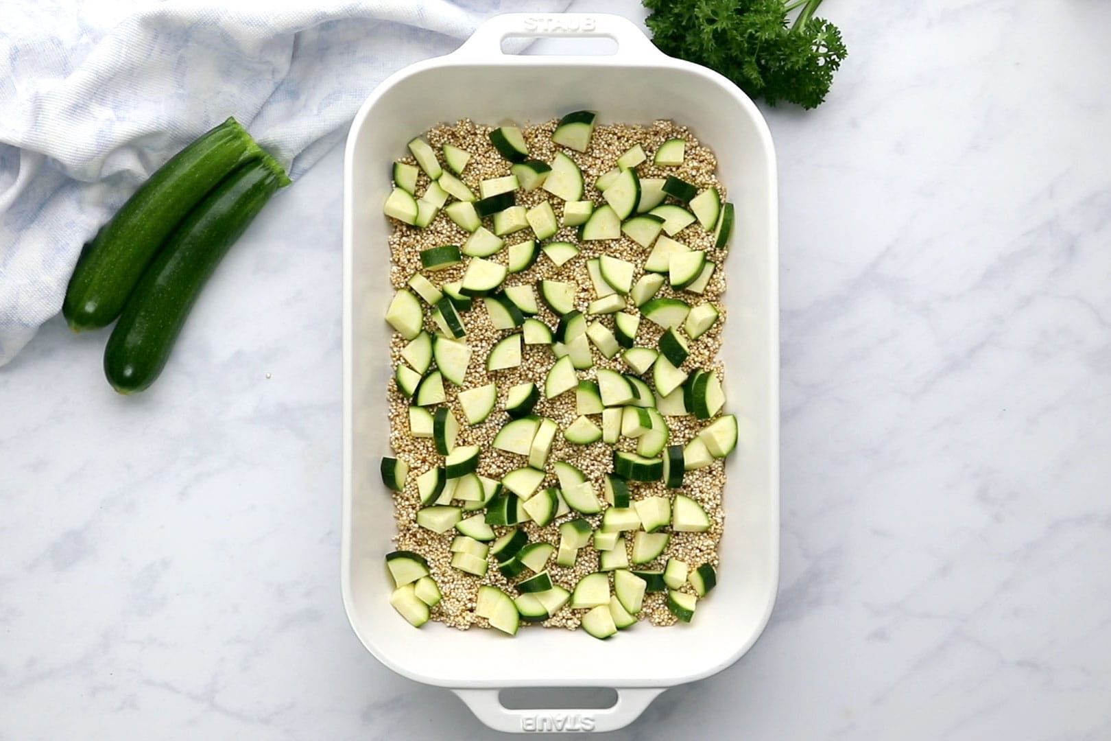Uncooked quinoa and sliced zucchini in a white casserole dish viewed from overhead
