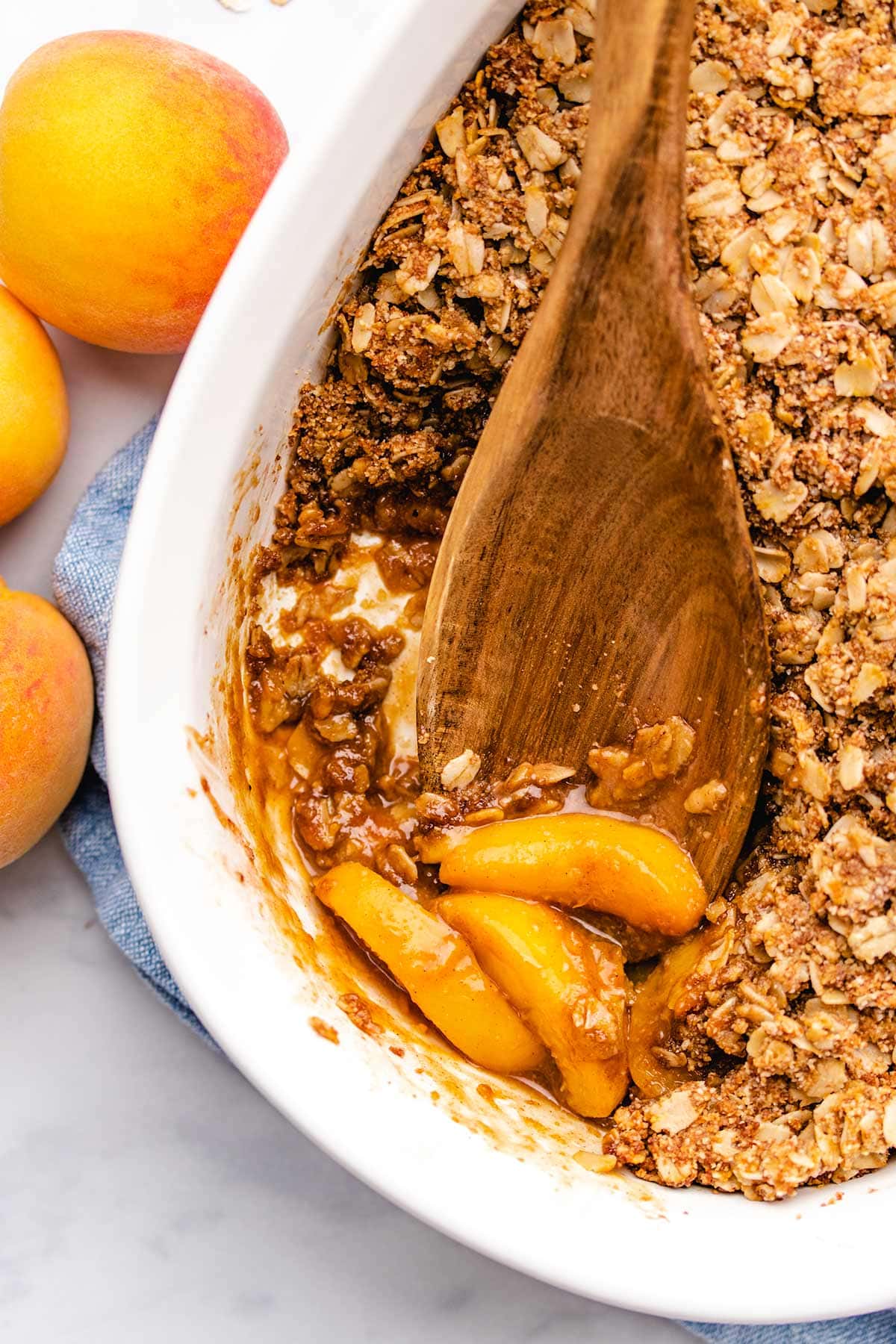 A wooden spoon taking a scoop of peach crisp from a white casserole dish