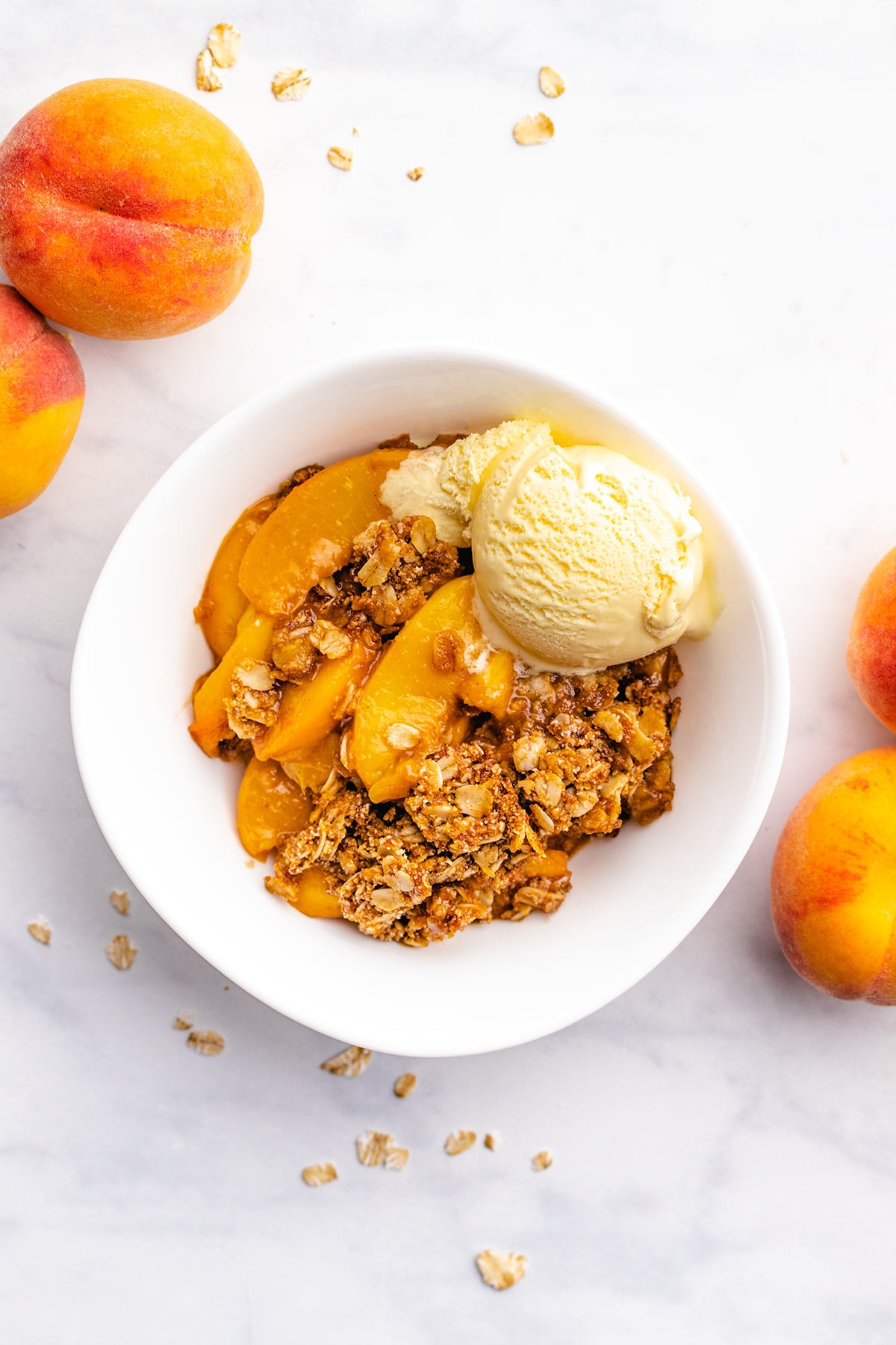 Peach Crisp in a white bowl with a scoop of vanilla ice cream surrounded by whole peaches