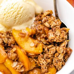 Peach Crisp in a white bowl with a scoop of vanilla ice cream and a spoon tucked in