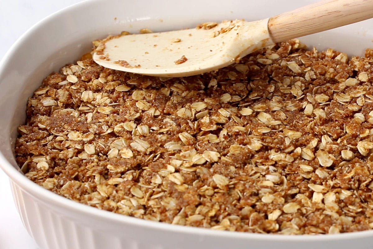 Oat and almond crisp mixture in a white casserole dish getting smoothed out with a spatula