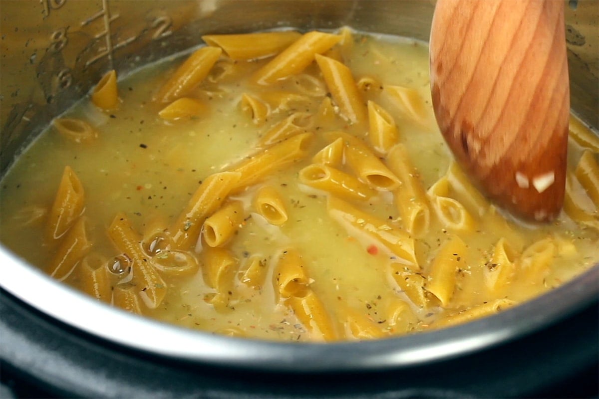 Using a wooden spoon to make an even layer of penne submerged in broth in an Instant Pot.