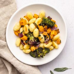 roasted gnocchi and vegetables on a white plate next to a brown linen and sage leaves