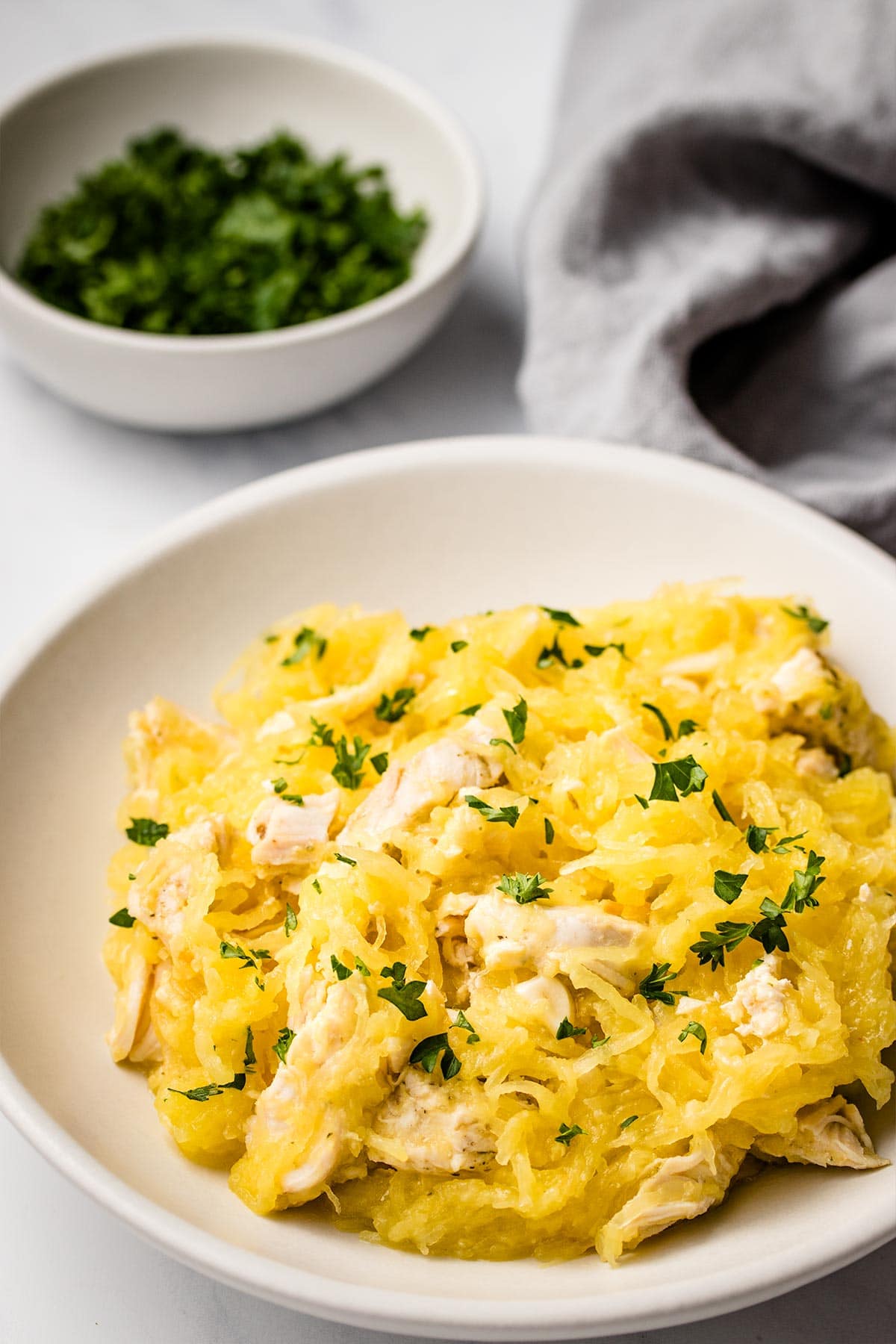 Spaghetti squash mixed with sliced chicken garnished with fresh parsley on a white plate, next to a bowl of parsley and a grey linen.