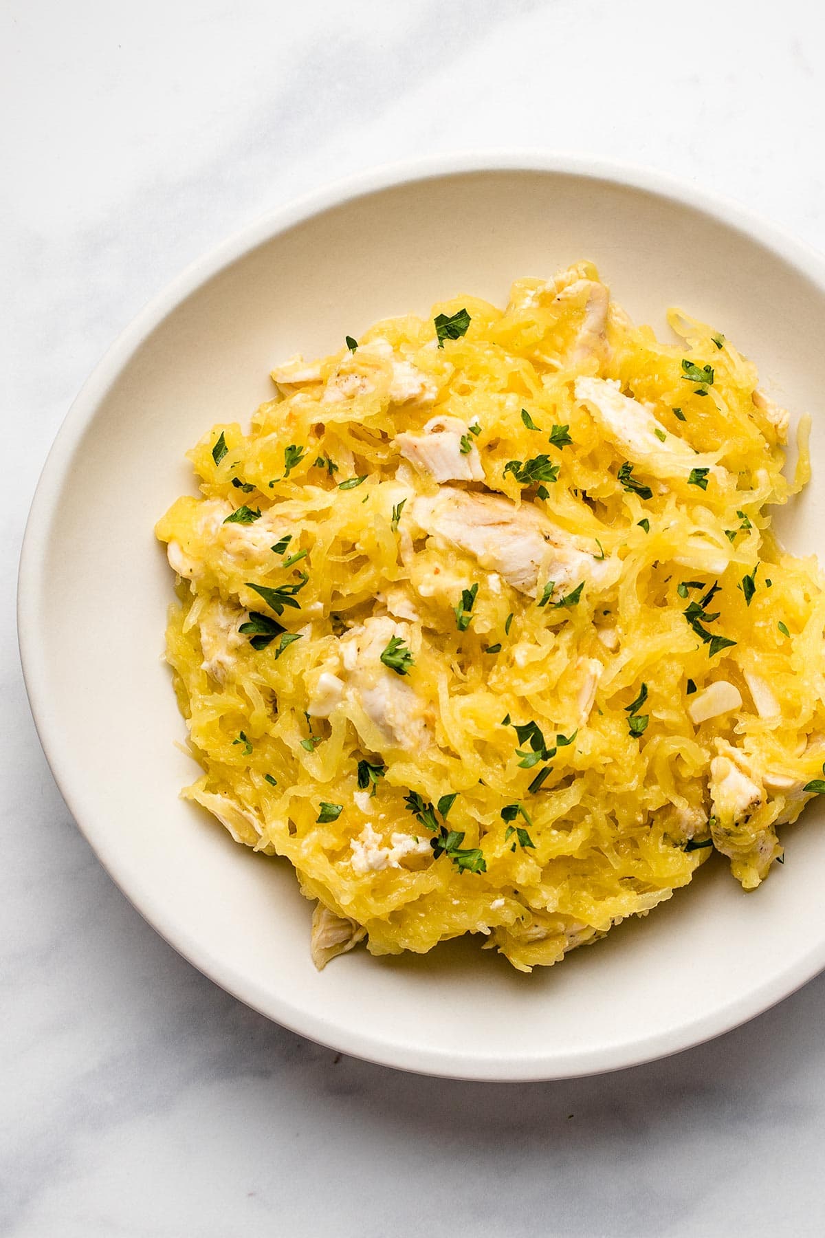 Spaghetti squash mixed with sliced chicken garnished with fresh parsley on a white plate viewed from overhead.