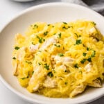 Spaghetti squash mixed with sliced chicken garnished with fresh parsley on a white plate.