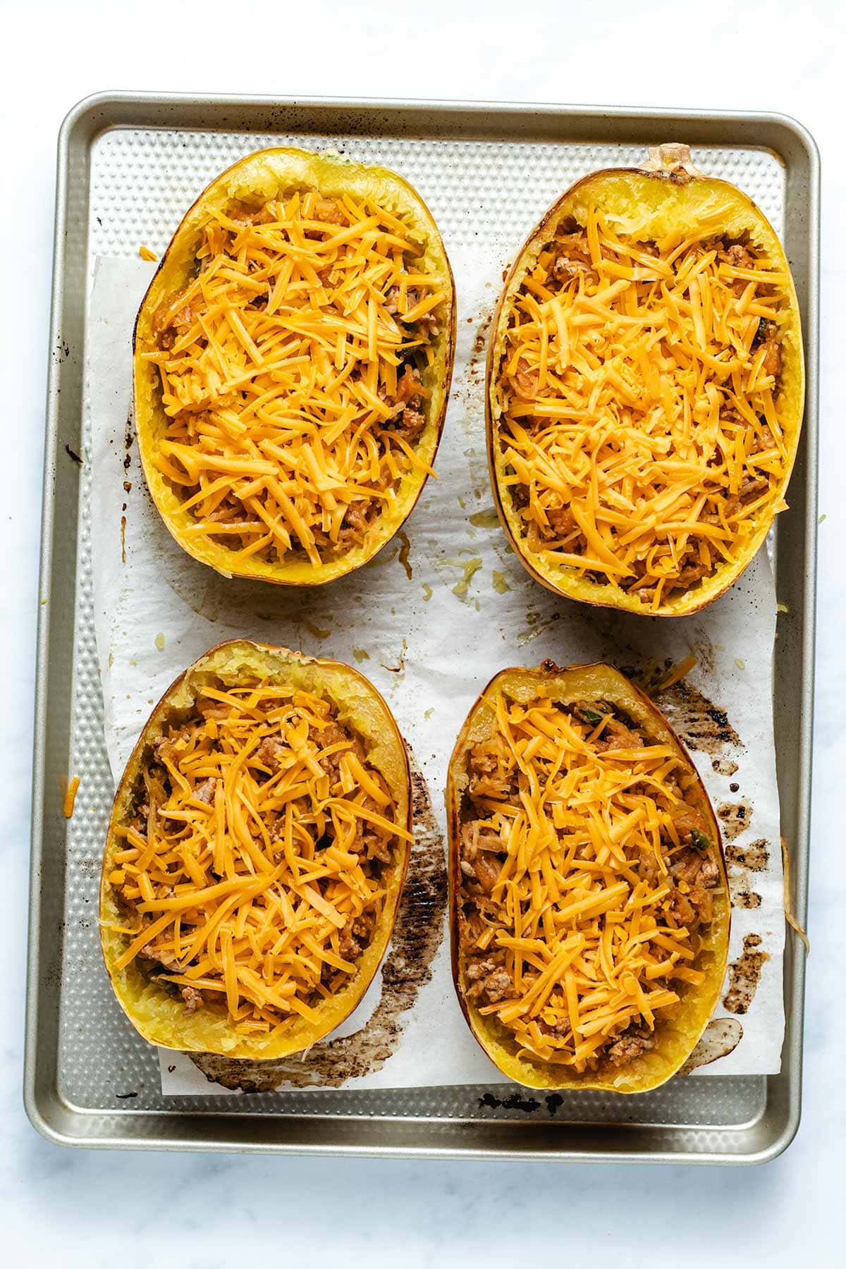 Four taco stuffed spaghetti squash halves on a sheet pan with grated cheese added on top before melting.