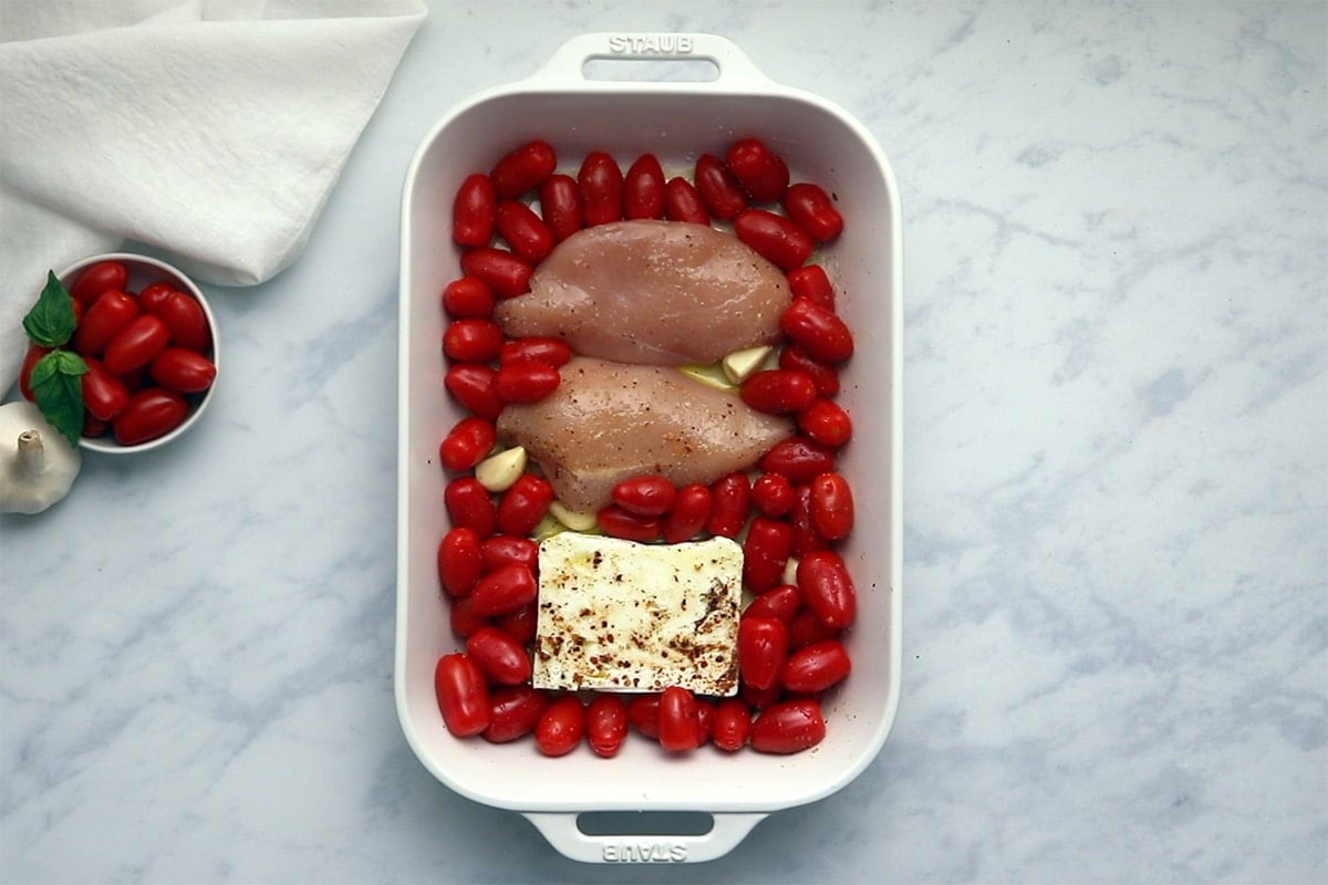 Uncooked chicken breasts, cherry tomatoes, feta cheese and garlic in a white casserole dish.