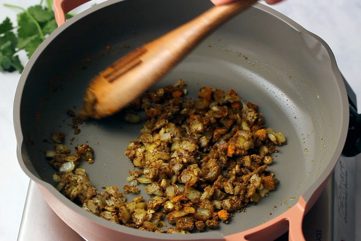 Mixing spices into aromatics in a pink and grey frying pan.