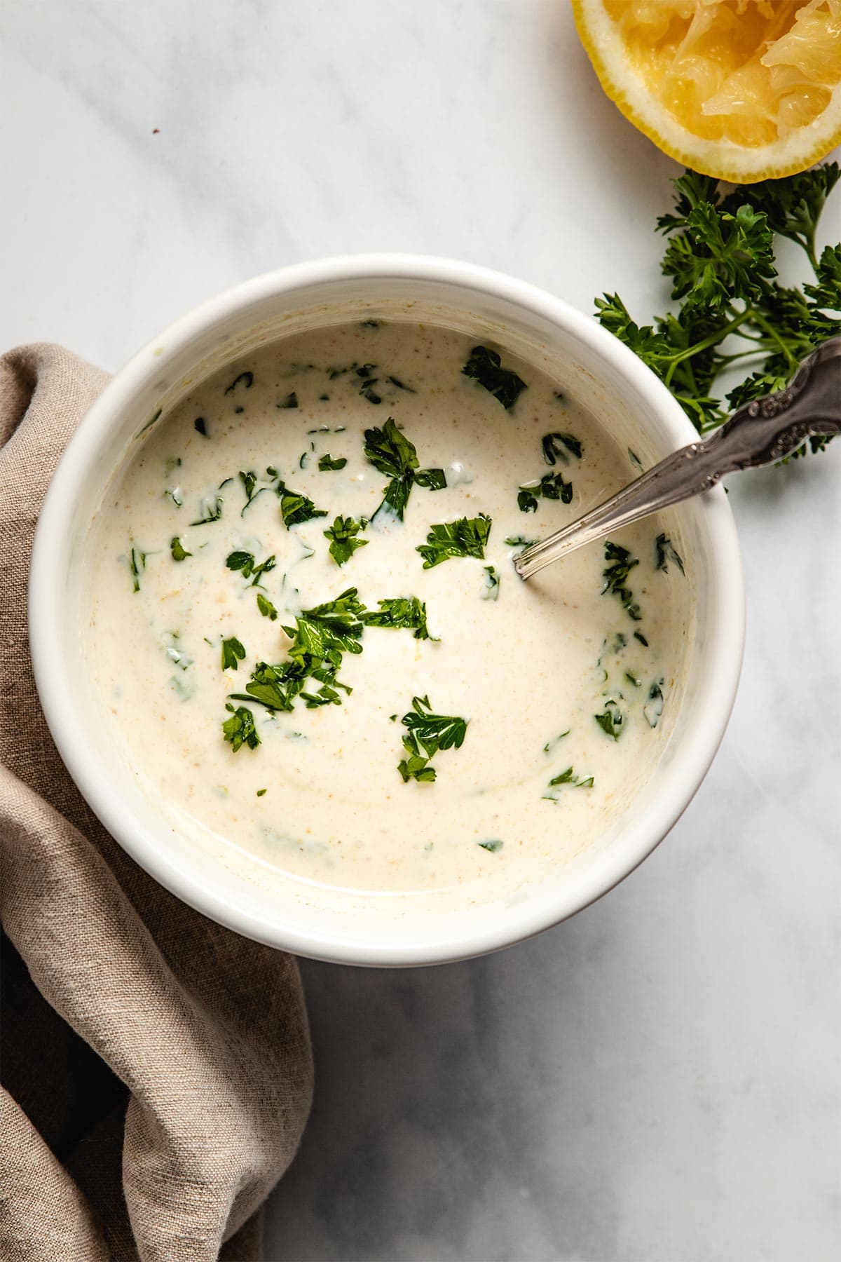 Lemon yogurt sauce in a white bowl garnished with parsley next to a brown linen, parsley and a squeezed lemon half.
