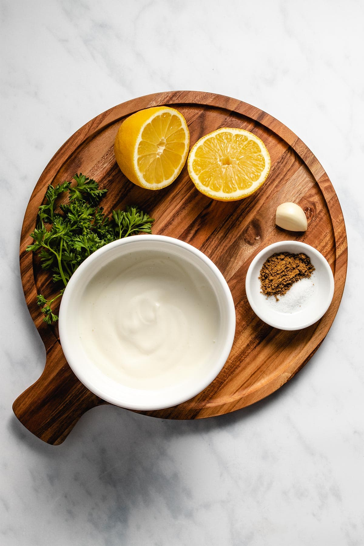 Ingredients for lemon yogurt sauce on a round wooden serving board viewed from overhead.