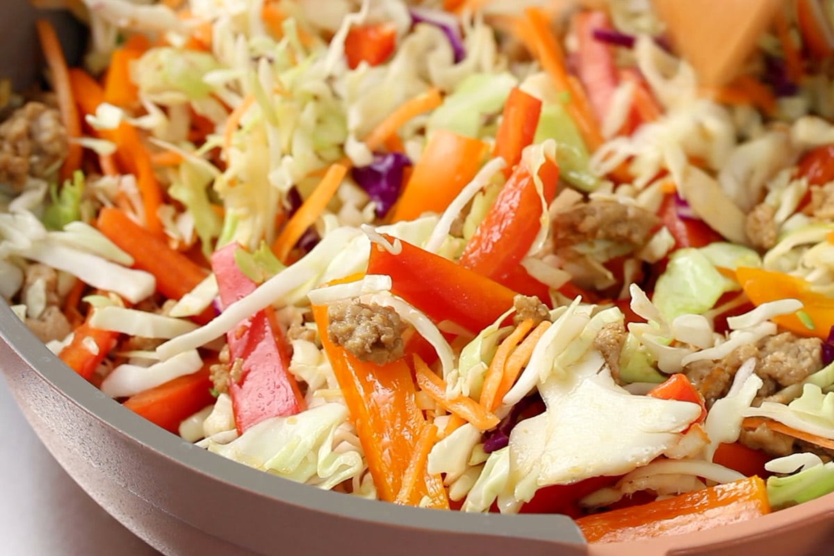 Coleslaw mix, bell peppers and chicken being cooked together in a pink and grey frying pan.