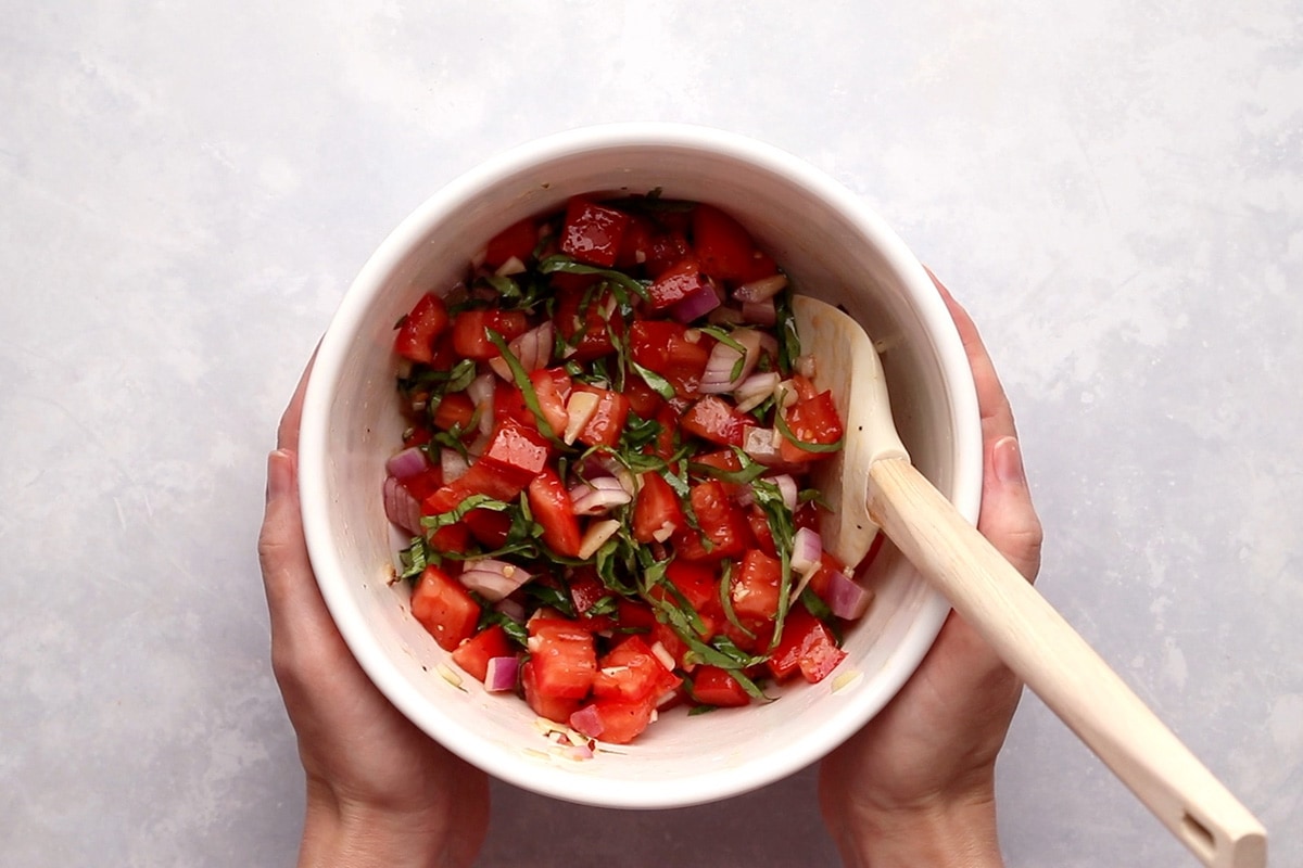 Bruschetta topping in a white bowl with a white spatula, being held by hands, viewed from overhead.