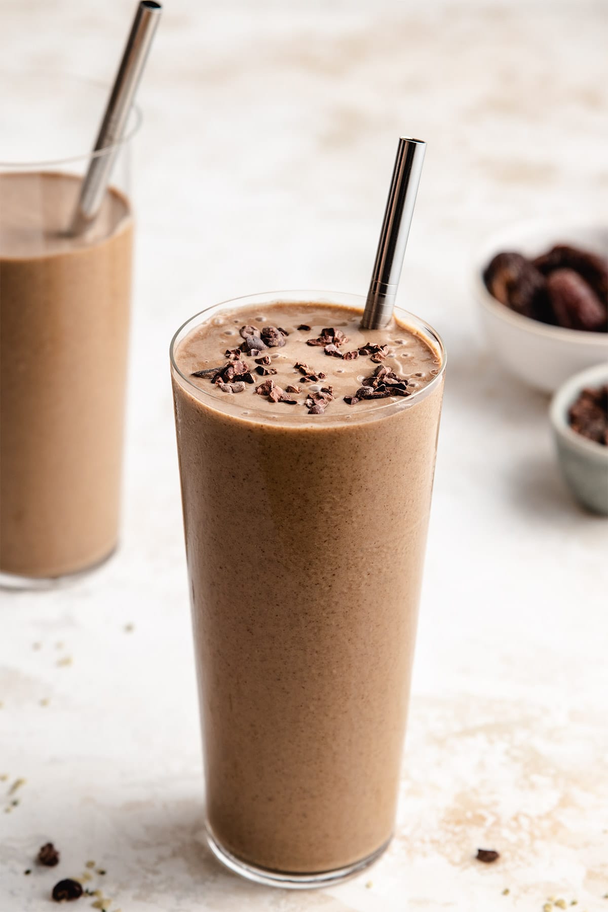 A coffee protein smoothie garnished with cacao nibs in a tall clear glass with a silver straw.