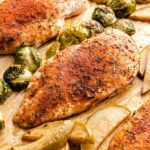 Roasted chicken breast with brown sugar seasoning, surrounded by apple slices and Brussels sprouts on a parchment-lined sheet pan.