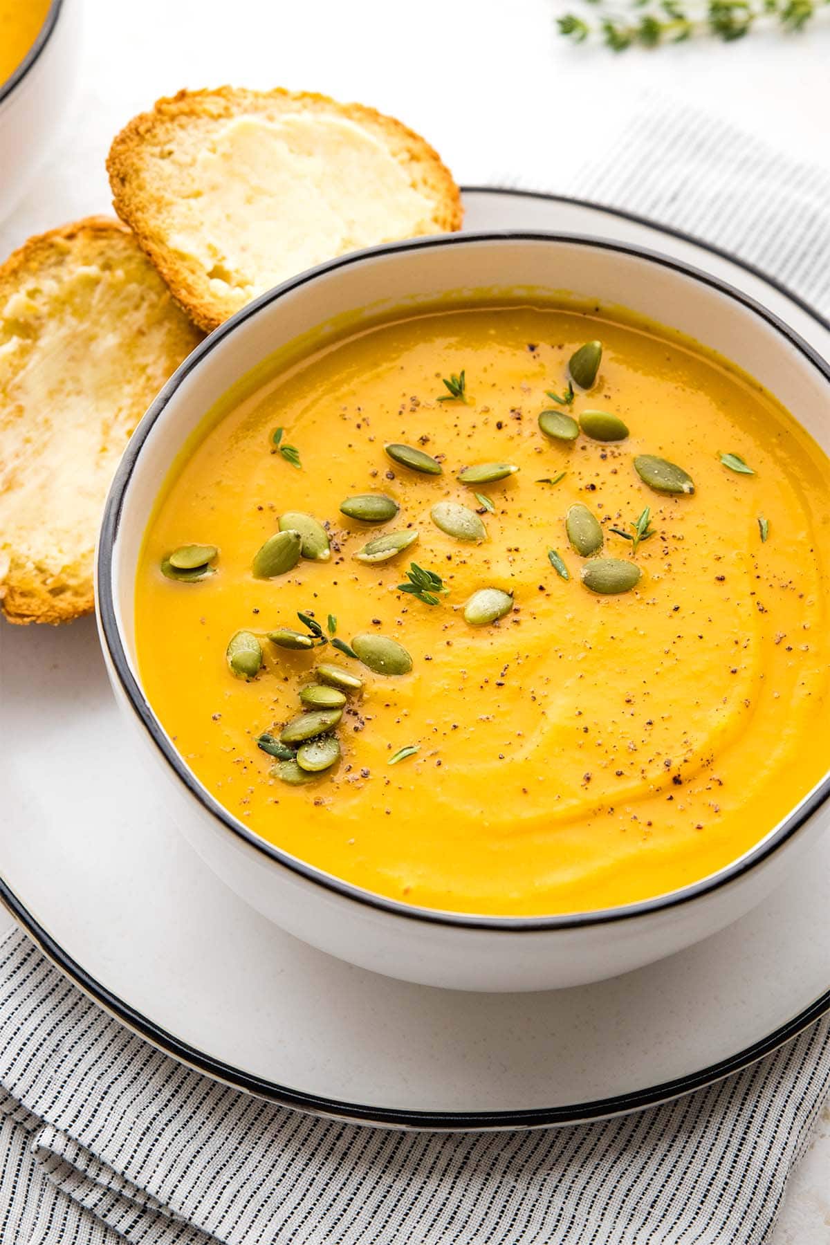 Butternut squash soup in a white bowl with a black rim, garnished with pumpkin seeds, thyme and black pepper, served with two buttered baguette slices.