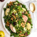 Kale and Brussels Sprouts Salad on a shallow, white oval serving dish, next to apples, pepitas, dried cranberries and serving spoons.
