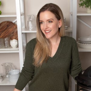 Portrait of Laura Lawless in a green sweater standing in front of white shelving filled with food photography props.