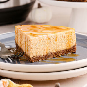 A slice of pumpkin cheesecake drizzled with caramel sauce, on a white and grey plate.