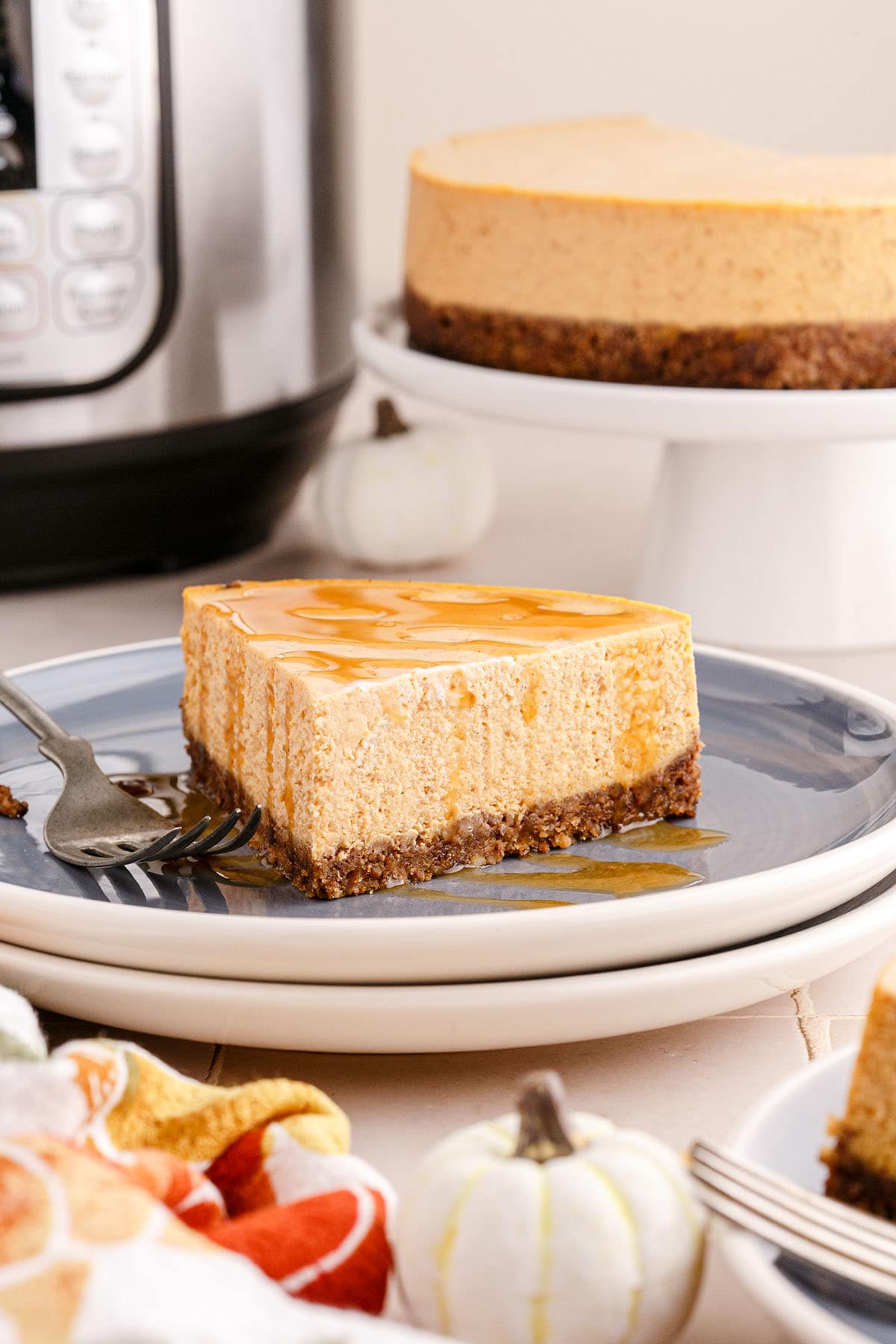 A slice of pumpkin cheesecake drizzled with caramel sauce, on a white and grey plate, in front of a cake stand and an Instant Pot.