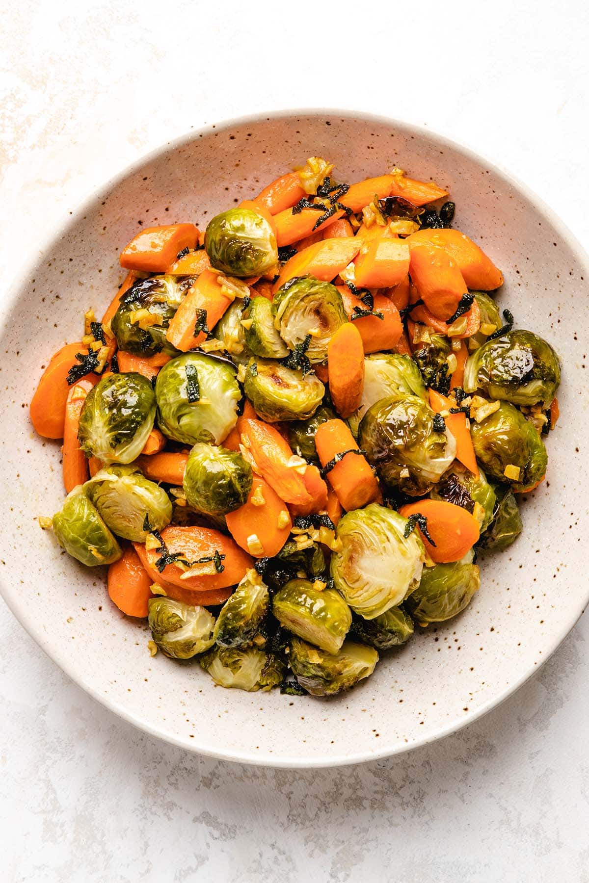 Roasted Brussels sprouts and carrots tossed with brown butter sage in a beige speckled bowl.