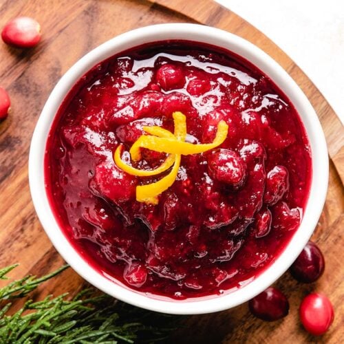 Cranberry sauce garnished with orange peel curls in a white bowl placed on a wooden board with winter greens.