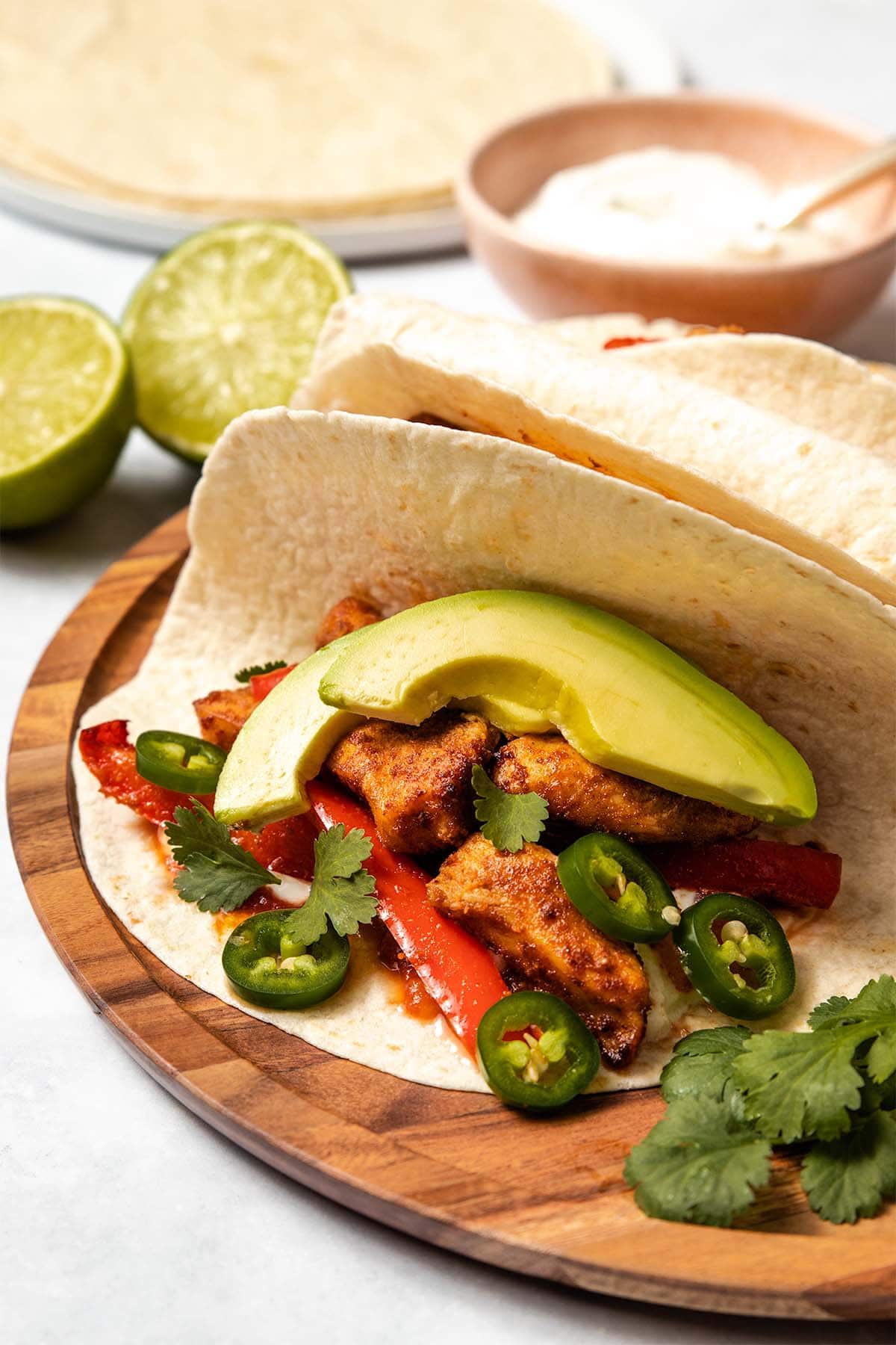 Chicken fajitas garnished with jalapeno, salsa, avocado and cilantro, served on a round wooden board.