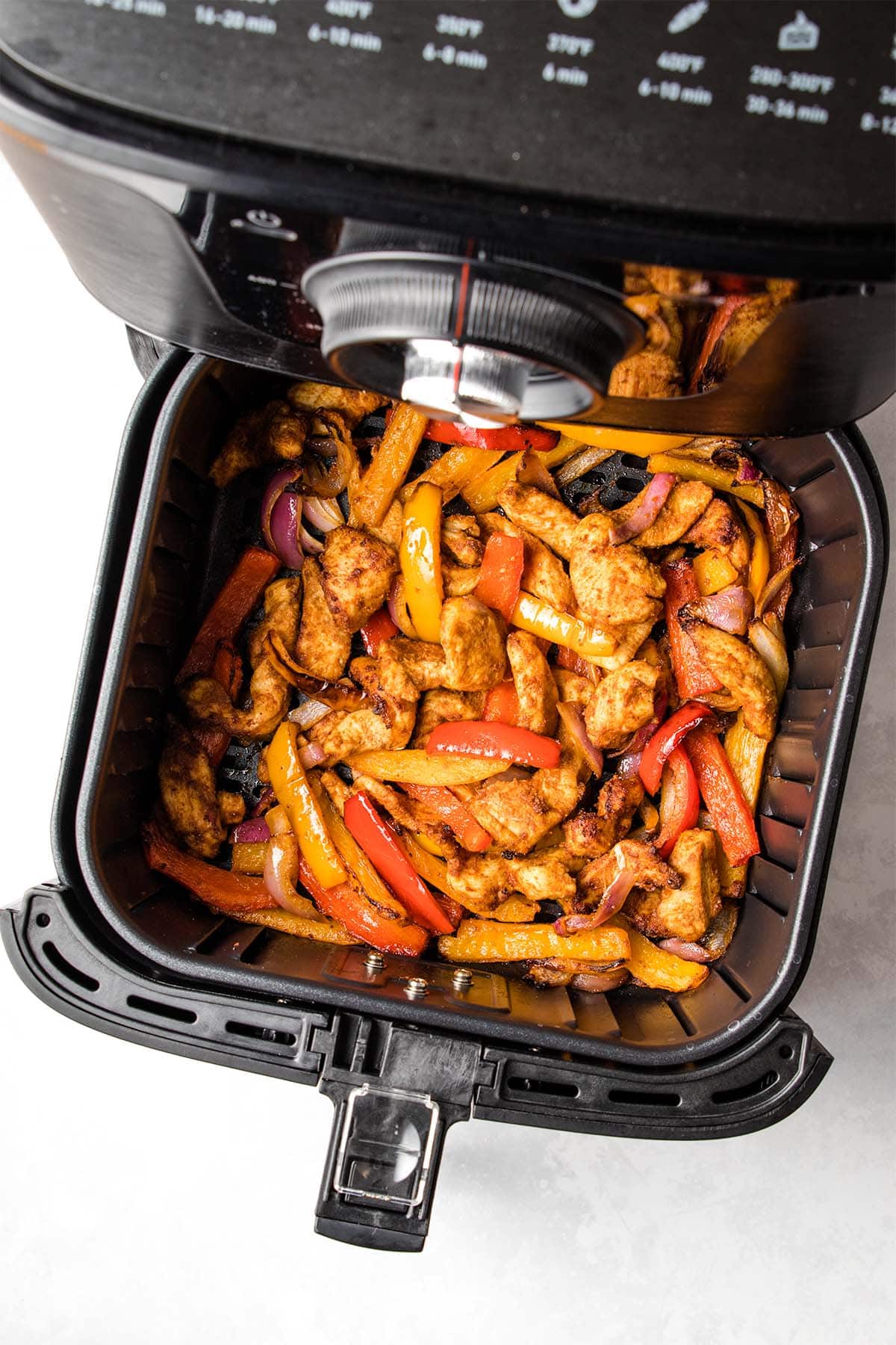 Cooked chicken strips, bell peppers and red onion coated in fajita seasoning in a black air fryer basket, viewed from overhead.